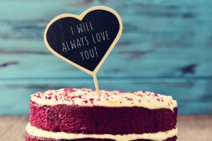 closeup of a red velvet cake topped with a heart-shaped chalkboard with the text I will always love you written in it, on a rustic wooden table